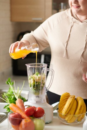 Photo for Smiling curvy young woman pouring orange juice in blender with cut fruits and vegetables - Royalty Free Image