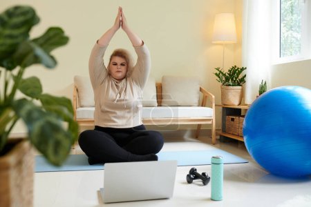 Foto de Mujer joven sentada en posición de loto delante de la computadora portátil abierta y levantando las manos en gesto namaste sobre la cabeza - Imagen libre de derechos