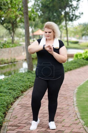 Téléchargez les photos : Plus-taille jeune femme vérifiant fitness tracker après avoir fait du jogging à l'extérieur le matin - en image libre de droit
