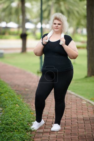 Téléchargez les photos : Portrait de jeune femme courbée en tenue de sport noire debout sur le chemin dans le parc après avoir couru - en image libre de droit