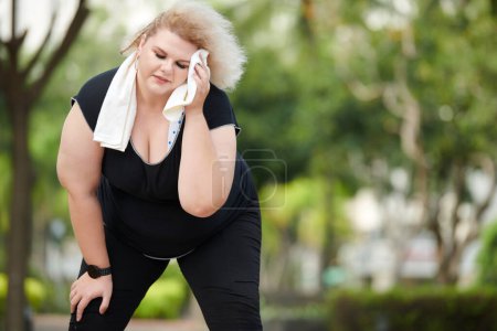 Foto de Mujer joven y sudorosa cansada de talla grande que se limpia la frente después de hacer ejercicio interno al aire libre - Imagen libre de derechos