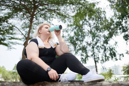 Photo for Curvy young woman sitting on stone curb in park and drinking electrolyte water after jogging in park - Royalty Free Image