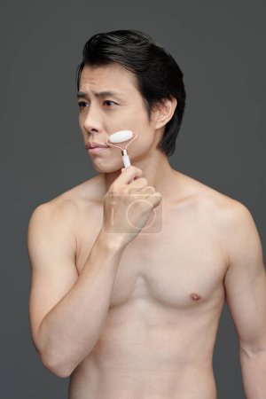 Foto de Retrato de estudio de hombre sin camisa masajeando la cara con rodillo de cuarzo después de la ducha - Imagen libre de derechos