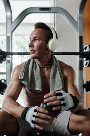 Foto de Deportista escuchando música en auriculares y agua potable cuando hace ejercicio en el gimnasio - Imagen libre de derechos