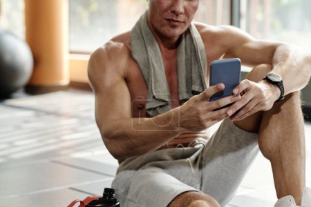 Téléchargez les photos : Fit homme assis sur le sol de la salle de gym et répondre aux messages texte - en image libre de droit
