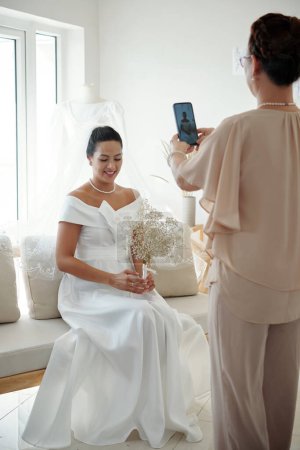 Téléchargez les photos : Souriant élégante jeune femme en robe de mariée assise sur le canapé dans l'atelier de couture - en image libre de droit
