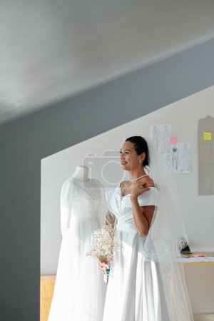 Foto de Hermosa novia alegre en vestido de novia blanco de pie en el salón con ramo de flores - Imagen libre de derechos