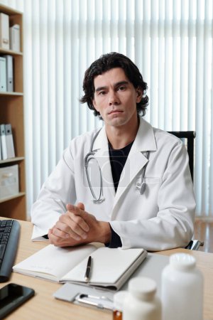 Téléchargez les photos : Portrait d'un médecin généraliste sérieux en manteau assis sur la table dans un cabinet médical - en image libre de droit