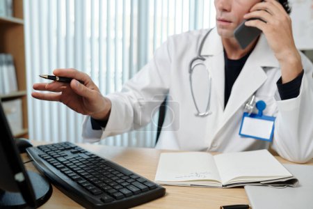 Téléchargez les photos : Médecin analysant les résultats des tests médicaux à l'écran de l'ordinateur lorsqu'il parle au téléphone avec le patient - en image libre de droit