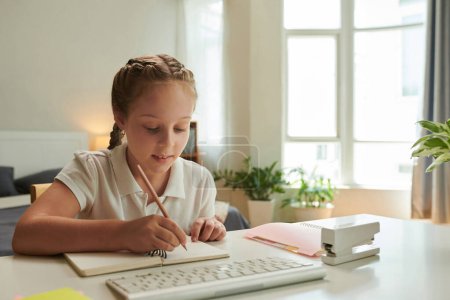 Foto de Sonriente niña preadolescente haciendo deberes en su escritorio, escribiendo en un libro de texto - Imagen libre de derechos
