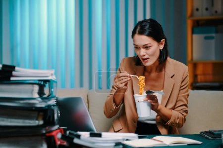 Téléchargez les photos : Femme d'affaires affamée manger nouilles instantanées en regardant la présentation sur ordinateur portable au bureau tard dans la nuit - en image libre de droit