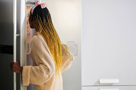 Foto de Mujer joven hambrienta abriendo refrigerador para tomar algo de comer - Imagen libre de derechos