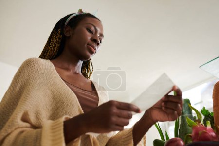 Foto de Mujer seria comprobando recibo de pago después de recibir comestibles entregados a su casa - Imagen libre de derechos