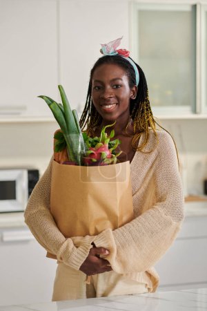 Photo for Portrait of happy Black woman standing in kitchen with paper bag of fresh groceries - Royalty Free Image