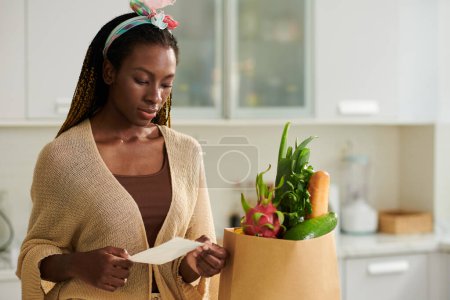 Foto de Mujer joven revisando su lista de compras después de recibir comestibles entregados a su casa - Imagen libre de derechos