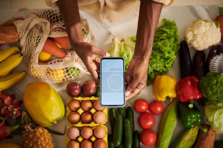 Photo for Hands of Black woman holding smartphone with empty screen over kitchen table with fresh fruits and vegetables - Royalty Free Image