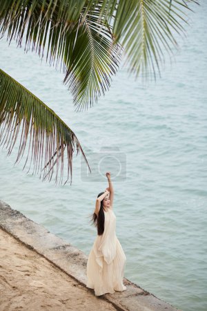 Photo for Young woman in long flowing beige dress enjoying fresh sea breeze on coast - Royalty Free Image