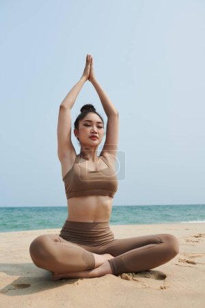 Foto de Mujer joven sentada en posición de loto cuando sus brazos sobre la cabeza al meditar en la playa - Imagen libre de derechos