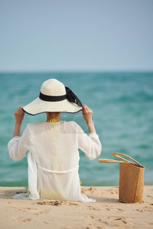Téléchargez les photos : Jeune femme en chapeau de paille assise sur une plage de sable, vue de dos - en image libre de droit