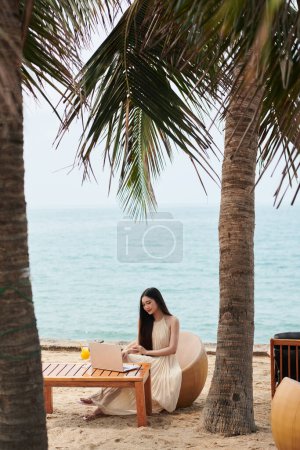 Téléchargez les photos : Jeune femme en robe d'été travaillant à table sur la plage tropicale - en image libre de droit
