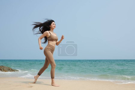 Téléchargez les photos : Jeune femme asiatique avec des cheveux longs jogging sur la plage le matin - en image libre de droit