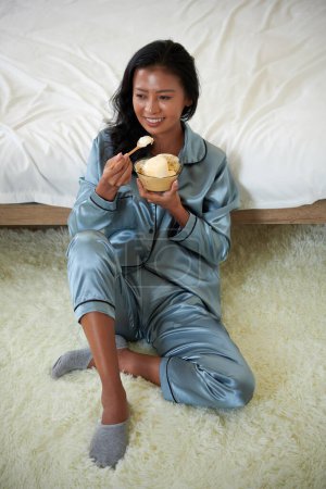 Photo for Smiling young woman in silk pajama sitting on floor and eating bowl of tasty vanila ice cream - Royalty Free Image