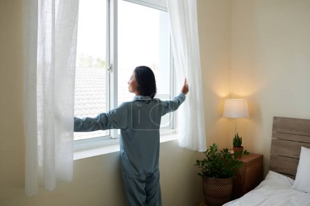 Téléchargez les photos : Jeune femme souriante en pyjama ouvrant les rideaux et regardant dehors - en image libre de droit