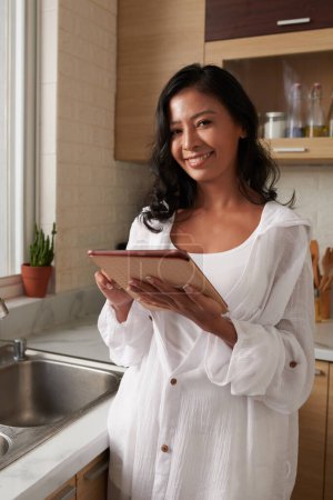 Photo for Portrait of smiling young woman standing in kitchen with tablet computer and looking at camera - Royalty Free Image
