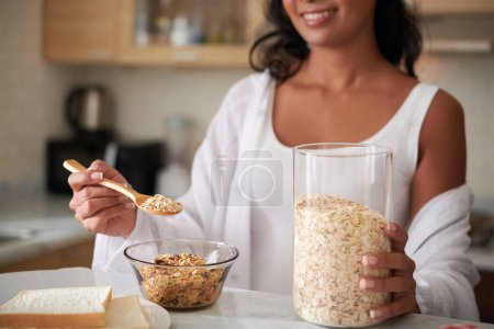 Photo for Young woman adding spoon of oat flakes in bowl when making tasty and healthy breakfast - Royalty Free Image
