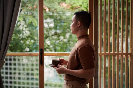 Foto de Joven pensativo de pie en el balcón, bebiendo café de la mañana y mirando a la calle - Imagen libre de derechos