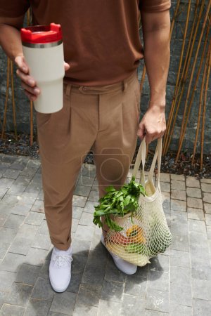 Photo for Young man holding reusable cup of coffee and mesh bag of fresh groceries - Royalty Free Image