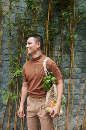 Téléchargez les photos : Portrait de jeune homme heureux tenant sac de maille d'épicerie fraîche qu'il a acheté au marché local - en image libre de droit