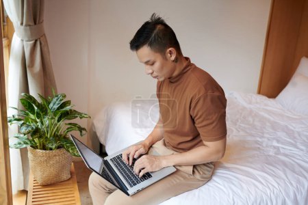 Téléchargez les photos : Concentré jeune homme assis sur le lit et vérifier le code de programmation sur l'écran d'ordinateur portable - en image libre de droit