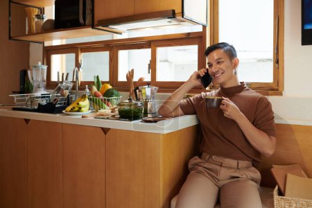 Téléchargez les photos : Jeune homme souriant assis au comptoir de la cuisine, buvant le café du matin et parlant au téléphone avec un ami - en image libre de droit