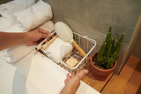 Téléchargez les photos : Mains d'homme mettant panier avec petite serviette, savon fait main et brosses à dents et brosse à cheveux en bambou écologiques biodégradables à côté de la baignoire - en image libre de droit
