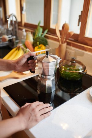 Téléchargez les photos : Mains de l'homme mettant la casserole de moka sur la cuisinière lors de la préparation du café du matin - en image libre de droit