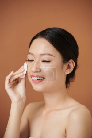 Téléchargez les photos : Souriant jeune femme essuyant visage avec coton tampon avec toner hydratant - en image libre de droit