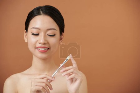 Téléchargez les photos : Souriant jeune femme prenant pinceau synthétique pour appliquer le maquillage - en image libre de droit