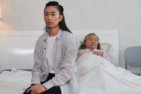 Foto de Mujer vietnamita grave casi llorando sentada en la cama de su madre anciana enferma - Imagen libre de derechos