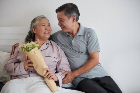 Foto de Mujer mayor feliz mirando al marido que trajo su ramo de flores en la cama - Imagen libre de derechos