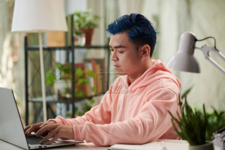 Téléchargez les photos : Sérieux jeune homme codant sur ordinateur portable à son bureau à la maison - en image libre de droit