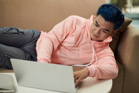 Photo for Lazy young man lying on couch and trying to code on laptop - Royalty Free Image