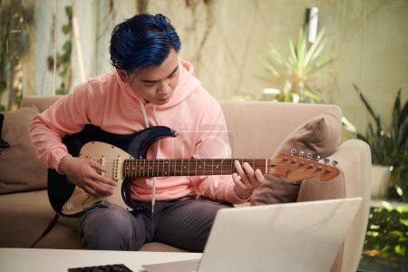 Foto de Hombre joven siguiendo tutorial en el ordenador portátil al aprender a tocar la guitarra en casa - Imagen libre de derechos