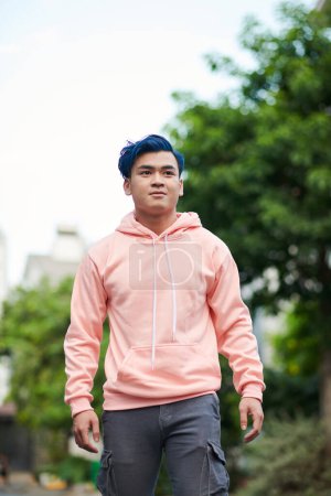 Photo for Positive teenage boy in salmon color hoodie walking in the street on summer day - Royalty Free Image