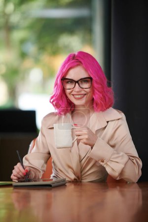 Téléchargez les photos : Portrait de heureuse jeune femme d'affaires aux cheveux roses buvant du café et signant des documents sur tablette - en image libre de droit