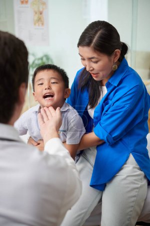 Téléchargez les photos : Médecin demandant à tout-petit garçon d'ouvrir la bouche afin qu'il puisse vérifier sa gorge - en image libre de droit