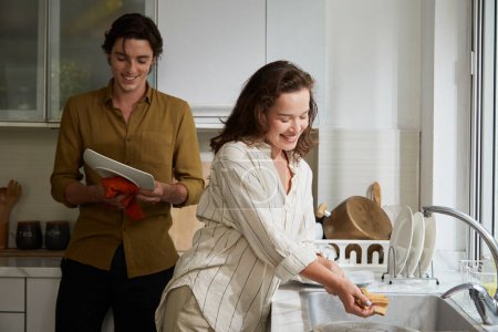 Téléchargez les photos : Femme souriante qui lave et monte la vaisselle lorsque son petit ami l'essuie avec un chiffon doux - en image libre de droit