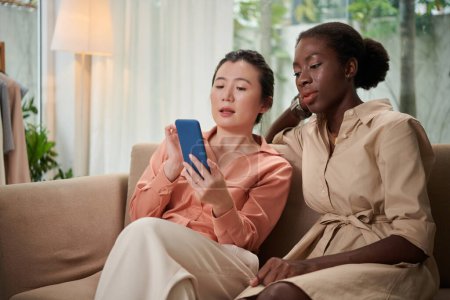 Téléchargez les photos : Femme assise sur le canapé avec un ami et montrant sa nouvelle application avec les tendances de la mode - en image libre de droit