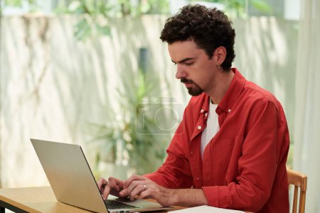 Téléchargez les photos : Homme en chemise rouge vif travaillant sur ordinateur portable dans un bureau ensoleillé - en image libre de droit