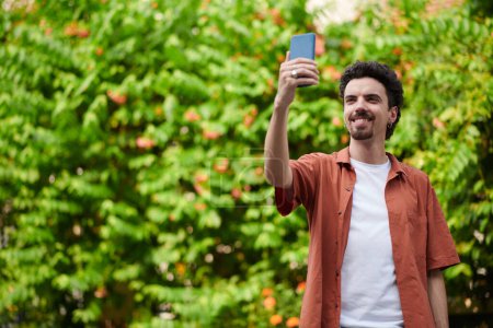 Photo for Smiling curly man taking selfie or video calling when walking outdoors - Royalty Free Image